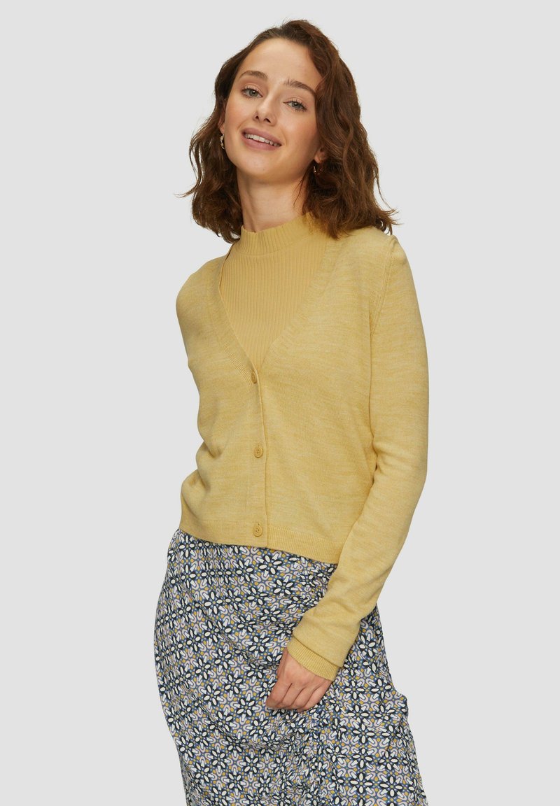 Gelber Cardigan mit V-Ausschnitt, Knopfverschluss und gerippter Textur, getragen über einem figurbetonten beigen Oberteil. Das Modell kombiniert ihn mit einem gemusterten Rock.