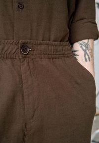 Brun linned jumpsuit med knaplukning foran, elastisk talje og sidelommer. Stoffet har en blød tekstur og et afslappet design.