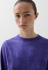 Lilla strikket sweater med ribbet hals og struktureret bølge-mønster foran. Blødt stof med en afslappet pasform.