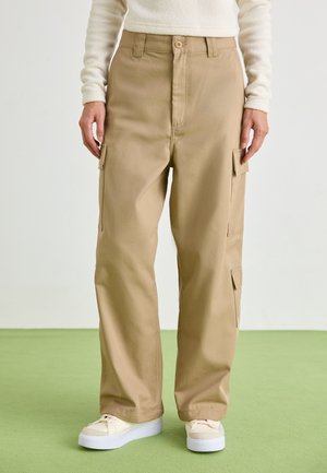 Persoon die beige wijde cargobroek met klepzakken en witte sneakers draagt, staand op een groene vloer tegen een effen achtergrond.