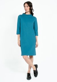 Tealfärgad klänning med avspänd passform, halterneck, trekvartslånga ärmar och rak skärning, tillsammans med svarta platåskor.