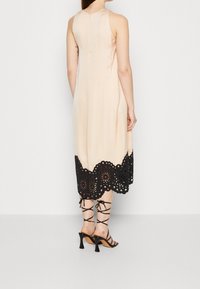 Robe sans manches beige avec un ourlet en dentelle noire comportant des découpes florales. Associée à des talons noirs à brides, mettant en valeur une silhouette ajustée.
