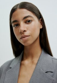 Grauer Blazer mit scharfen Revers, strukturiertem Stoff und einer klaren, maßgeschneiderten Passform. Das Model hat lange, glatte Haare und minimales Make-up.