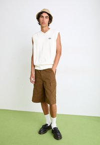 Hvit ermeløs genser, brune cargo-shorts, svarte platå-sko og hvite sokker. Bærer en beige bøttehatt med logo-detalj.