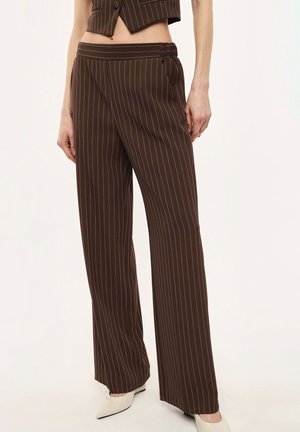 Pantaloni marroni a righe larghe abbinati a un gilet corto coordinato e scarpe bianche con punta appuntita, visti dalla vita ai piedi.