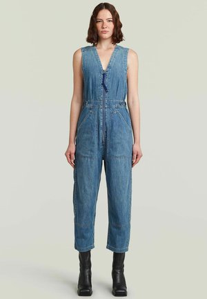 Frau mit schulterlangem Haar, die einen ärmellosen blauen Denim-Overall mit Frontreißverschluss und schwarzen Knöchelstiefeln trägt und vor einem schlichten Hintergrund steht.