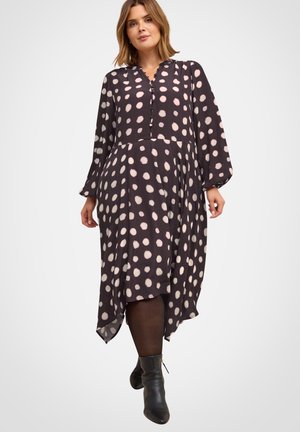 Frau in einem schwarzen Midikleid mit hellrosa Polka-Dots, langen Ärmeln und schwarzen Stiefeletten, die vor einem weißen Hintergrund steht.