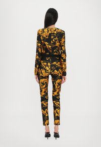Abito nero con motivo barocco dorato, design aderente, maniche lunghe e pantaloni slim. Scarpe col tacco alto nere e lucide completano il look.