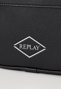 Crna torba od teksturirane umjetne kože s srebrnim logotipom u obliku dijamanta s natpisom "REPLAY" u središtu.
