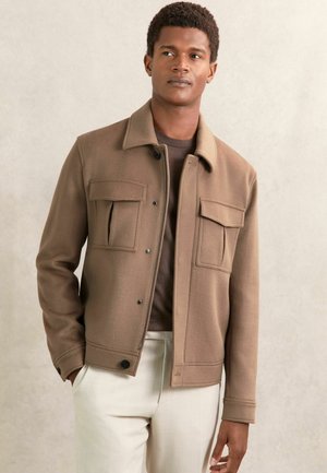 Braune Fleecejacke mit Kragen, zwei Brusttaschen und Druckknopfverschlüssen. Glatte Textur, leicht oversized Schnitt, kombiniert mit hellen Hosen.