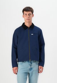 Chaqueta azul marina con un cuello de pana negro, cremallera frontal y bolsillo en el pecho; fabricada con un tejido resistente; cuenta con dos bolsillos laterales.