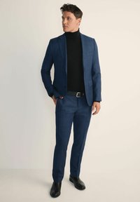 Granatowy garnitur w kratę o dopasowanym kroju, noszony na czarnym golfie. Gładka tekstura, zestawiony z czarnymi eleganckimi butami. Do zestawu dołączony prosty czarny pasek.