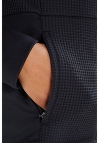 Main dans la poche zippée d'une veste noire texturée montrant les détails du tissu et les coutures.