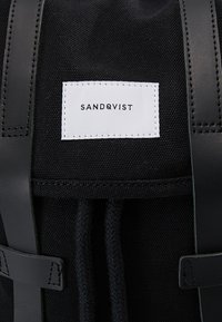 Sac à dos en tissu noir avec des bretelles en cuir noir et une étiquette blanche affichant le nom de la marque Sandqvist en lettres noires.