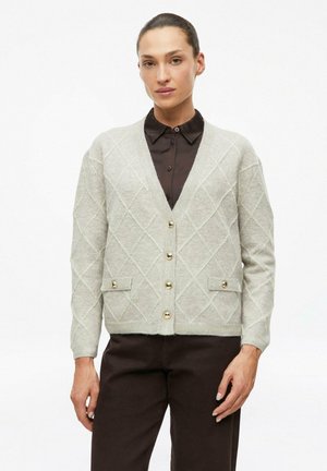 Frau trägt eine hellgraue, diamantmusterte Strickjacke mit goldenen Knöpfen über einem schwarzen Hemd mit Kragen und dunklen Hosen vor weißem Hintergrund.