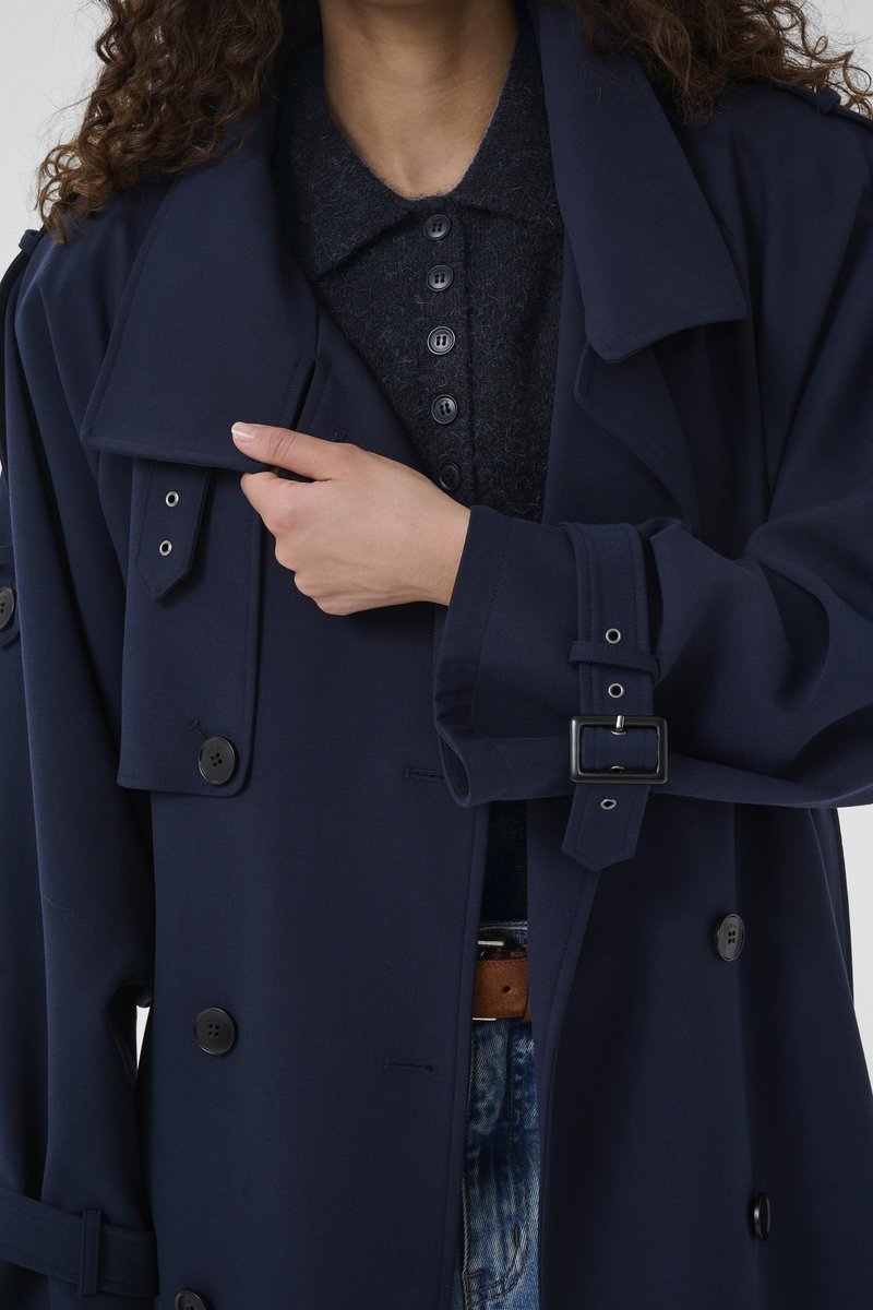 Trench-coat bleu marine avec de grands boutons noirs, un large col et une taille ceinturée. Le tissu semble lisse avec un design structuré.