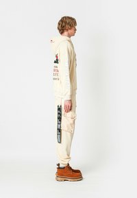 Hombre rubio con sudadera crema y pantalones con estampados gráficos y detalle de etiqueta negra, vistiendo botas gruesas marrones, de pie de lado sobre un fondo blanco.