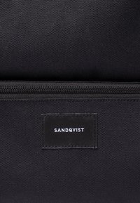 Gros plan d'un sac en tissu noir avec fermeture éclair visible et étiquette rectangulaire cousue affichant le nom de la marque Sandqvist en lettres blanches.