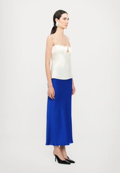 Camiseta de satén blanca con bordes en forma de escalloped, combinada con una falda midi azul y tacones de charol negros. La modelo está de perfil frente a un fondo sencillo.
