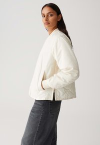 Veste bomber crème matelassée avec des poignets côtelés, des poches latérales et une fermeture éclair, associée à un jean gris foncé sur un fond neutre.