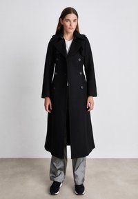 Lång svart rock i ull med dubbelknäppning, stora knappar, vid krage och sidofickor, bärs över grå byxor och svarta skor.