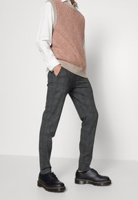 Gilet en tricot à motifs dans des teintes rouille et crème, porté par-dessus une chemise blanche. Pantalon à carreaux gris avec une coupe ajustée et chaussures noires à lacets avec une semelle épaisse.