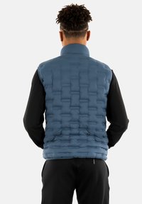 Gilet imbottito di colore blu scuro, caratterizzato da una texture trapuntata e un collo alto. Indossato sopra una maglietta nera a maniche lunghe. Pantaloni neri visibili.