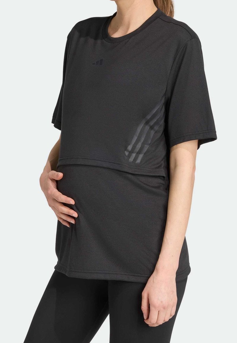 Personne enceinte en tenue de sport noire à manches courtes, tenant son ventre de la main gauche, debout sur un fond clair uni.