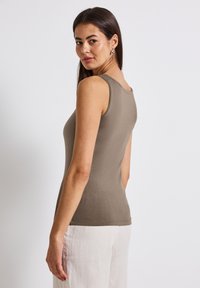 Khaki ærmeløs top med rund halsudskæring, tætsiddende design og glat bomuldsstof. Modellen bærer den med lysfarvede linnedbukser.