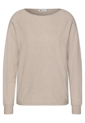 Femme aux cheveux bruns mi-longs souriante, portant un pull beige à manches longues et tenant un sac à bandoulière marron.