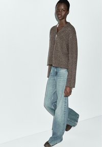 Brązowy prążkowany, krótki kardigan z dekoltem w serek, zestawiony z jasnoniebieskimi, szerokimi dżinsami. Strój cechuje się luźnym krojem i fakturowaną tkaniną.