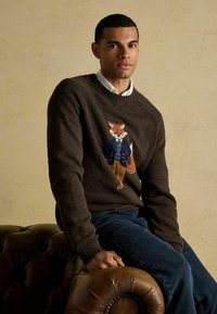 Young man sitting on brown leather sofa, wearing dark brown sweater with fox in jacket and trousers design, and blue jeans.