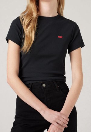 Femme portant un t-shirt noir ajusté Levi’s avec un petit logo rouge sur la poitrine, associé à un jean noir taille haute.