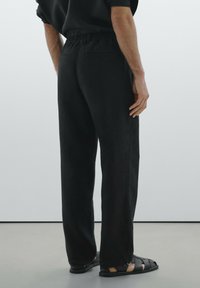 Pantalons noirs amples avec une taille élastique, dotés de poches latérales et d'une texture lisse, associés à des sandales noires à brides.