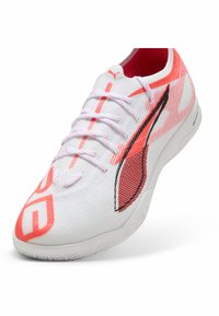 Chaussure de sport blanche avec un dessus en mesh respirant, accents orange, logo noir et semelle texturée. Design à lacets pour un ajustement sécurisé.