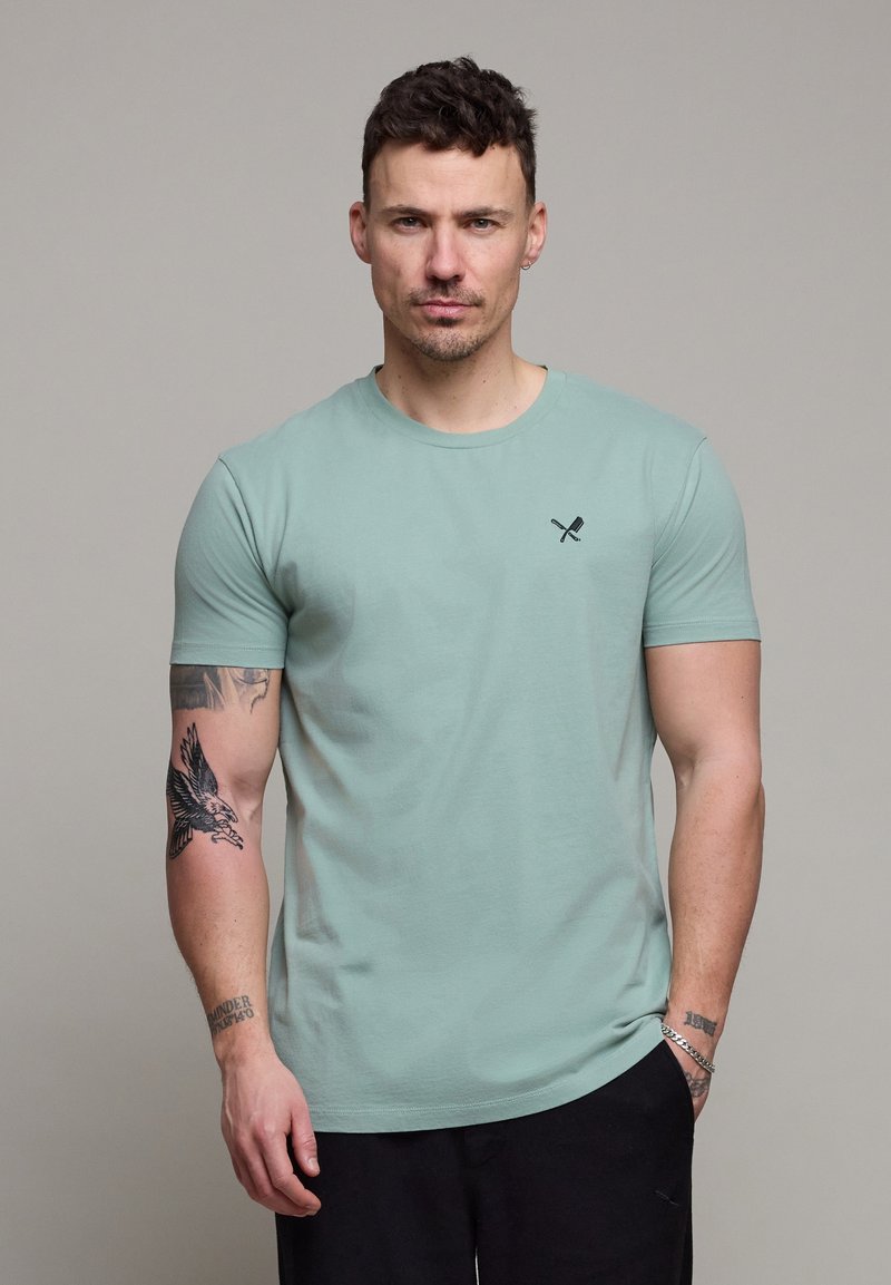 Homme aux cheveux courts et foncés portant un t-shirt vert clair avec un petit logo de cuillère et fourchette croisées, des tatouages sur le bras gauche et un pantalon noir.