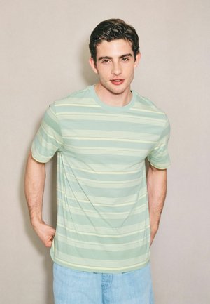 Junger Mann mit kurzen dunklen Haaren, der ein hellgrün-gelb gestreiftes T-Shirt und hellblaue Jeans vor einem einfarbigen Hintergrund trägt.