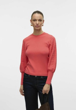 Koralfarvet strikket sweater med teksturerede puffærmer, rund halsudskæring og ribbet manchetter, parret med mørke jeans og en brun taske.