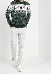 Grüner Strickpullover mit weißen Schneeflockenmustern und festlichen Designs, kombiniert mit hellgrauen Hosen und weißen Sneakern.