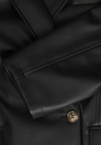 Manche de veste en cuir noire repliée sur le devant avec des coutures visibles et un unique bouton rond marron.