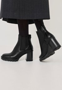 Černé kotníkové boty s lesklým koženým povrchem, elastickými bočními panely a robustním podpatkem. Texturovaná gumová podrážka s vzorkem pro lepší přilnavost.