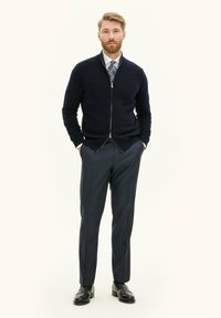 Cardigan zip-up blu navy sopra una camicia bianca, abbinato a pantaloni grigi scuri su misura e scarpe nere; texture liscia e design aderente.