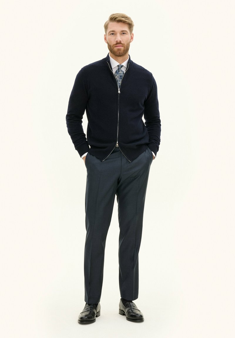 Cardigan zip-up blu navy sopra una camicia bianca, abbinato a pantaloni grigi scuri su misura e scarpe nere; texture liscia e design aderente.