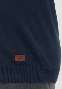 Jersey de color azul marino con dobladillo acanalado; presenta una etiqueta de cuero marrón etiquetada "BLEND" cerca de la parte inferior, mostrando un detallado bordado.