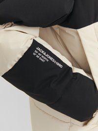Pufferjack met een zwart en crèmekleurig kleurblokontwerp, gemaakt van glad, gewatteerd materiaal. Bevat een logo met coördinaten op de mouw.