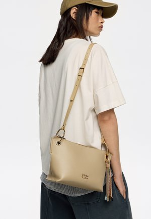 Bolso cruzado de cuero beige con un acabado texturizado, correa ajustable y herrajes dorados, que presenta un acento de borlas coloridas y una marca.