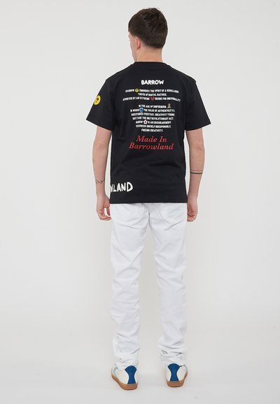 T-shirt nera con testo sulla schiena, caratterizzata da vari font e colori. Indossata con pantaloni bianchi e calzature con accenti blu e crema.