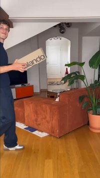 Kartono pakuotė su "Zalando" logotipu, laikoma žmogaus. Vidaus aplinkoje matyti tekstūruota oranžinė velveto sofa ir augalai.