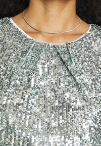 Haut en sequins argentés avec un col froncé, présentant une surface texturée. Accompagné d'un fin collier en chaîne argentée.