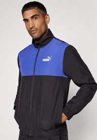 Schwarze und blaue Trainingsjacke mit Reißverschlussfront, elastischen Bündchen und Bund. Mit einem weißen Puma-Logo auf der Brust und glatter Stofftextur.
